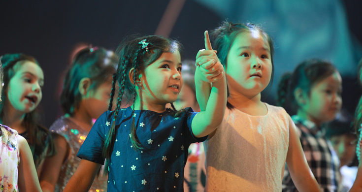 Two little girls on stage with one using the other girl's hand to show her something she is pointing to.
